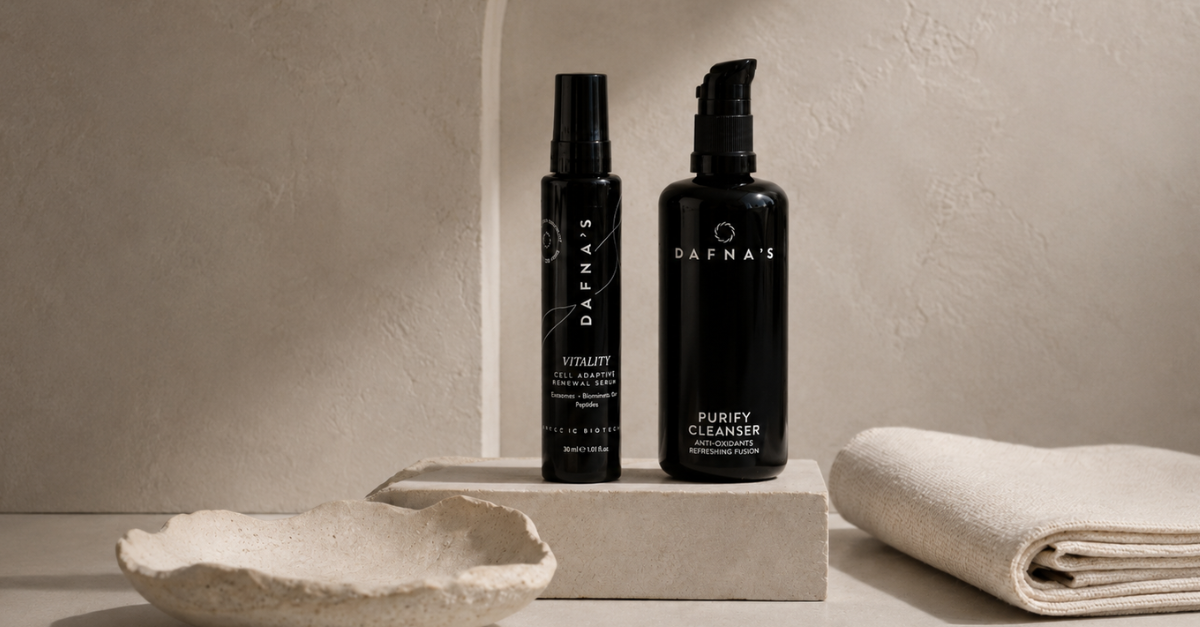 Two black skincare bottles from Dafna's, one labeled Vitality and one labeled Purify Cleanser, displayed on a stone pedestal with a folded cream towel and ceramic dish in a warm, minimalist setting.